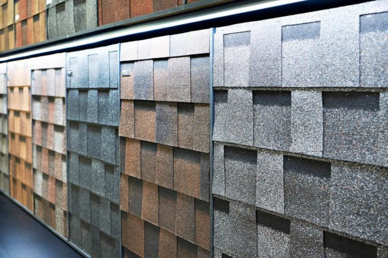 A display of various colored roof shingles, arranged in vertical panels, highlights the latest roofing trends. The textures range from smooth to rough, with colors including brown, gray, and reddish tones, all illuminated by overhead lighting.