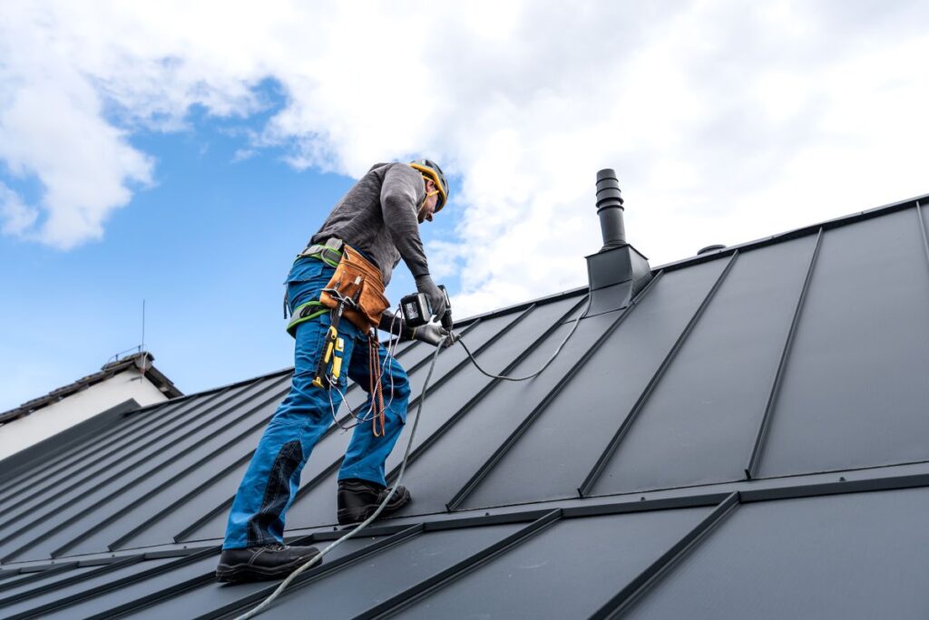 A worker wearing safety gear stands on a metal roof, using roofing tools and power tools for maintenance or installation. The sky is partly cloudy, and the worker is secured with a harness and rope.