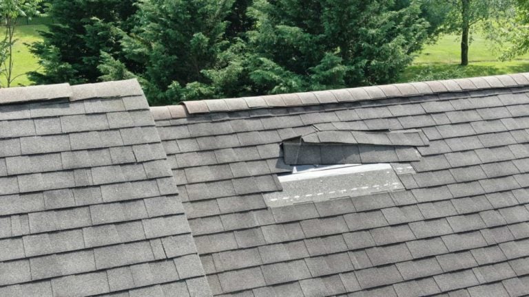A roof with dark shingles has a damaged section where blown off shingles have exposed the underlayment, leading to an urgent need for replacement. Green trees are visible in the background.
