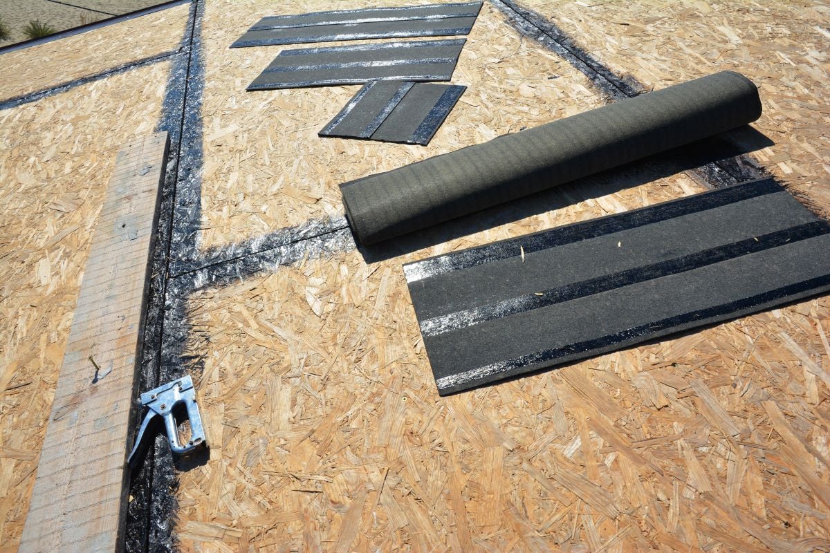 A roof under construction with black tar paper strips on plywood sheathing, roofing tools like a staple gun, and a roll of roofing material. Black adhesive lines mark where the material will be attached. Roofing Tools