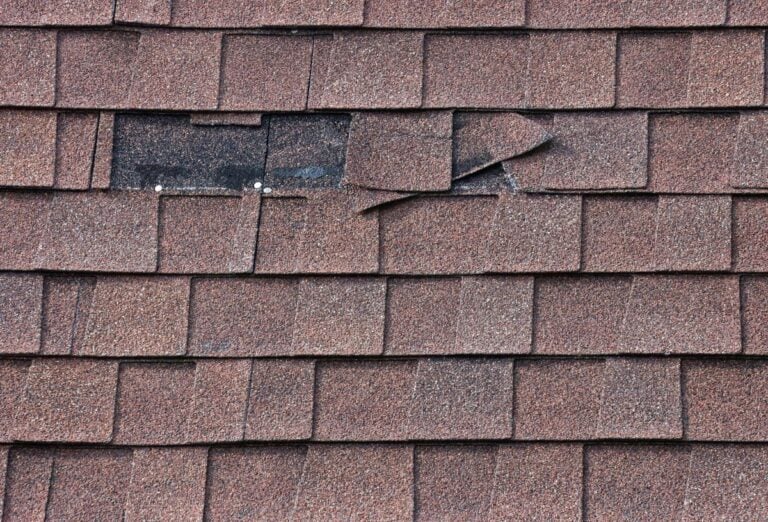 Close-up of a brown shingle roof with several damaged and misaligned shingles, exposing the black underlayment and roofing nails beneath—highlighting the need to replace blown off shingles.