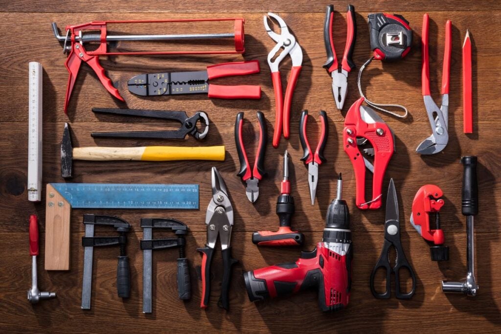 A variety of hand tools, including roofing tools like pliers, screwdrivers, clamps, tape measure, scissors, rulers, a hammer, wire cutters, and a red cordless drill are arranged neatly on a wooden surface.