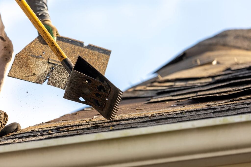 A person uses roofing tools to remove old, damaged shingles from a roof during a repair or replacement on a sunny day.