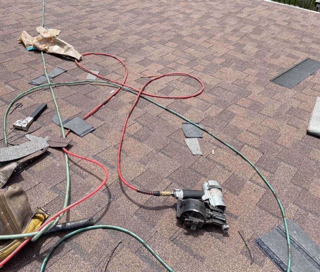 A close-up view of a roof under repair, featuring brown shingles, roofing tools like a pneumatic nail gun with air hoses, loose materials, and a brown tool bag on the roof surface.