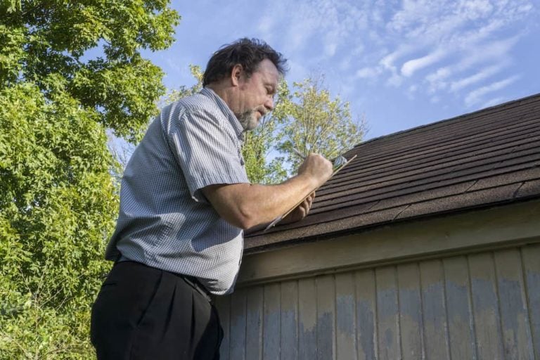 A person stands on a ladder inspecting a roof while writing on a clipboard. The scene is outdoors with green trees and a partly cloudy blue sky in the background.