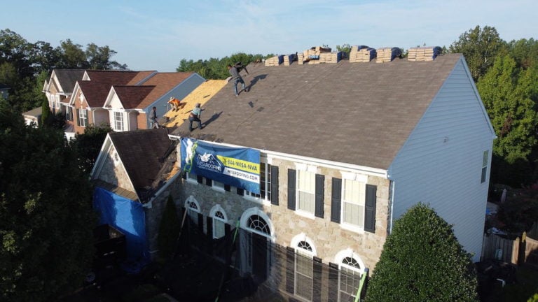 Workers are replacing the roof of a large suburban house. They are placing new shingles while bundles are stacked nearby. A sign is attached to the roof. Trees line the background, and neighboring houses are visible.
