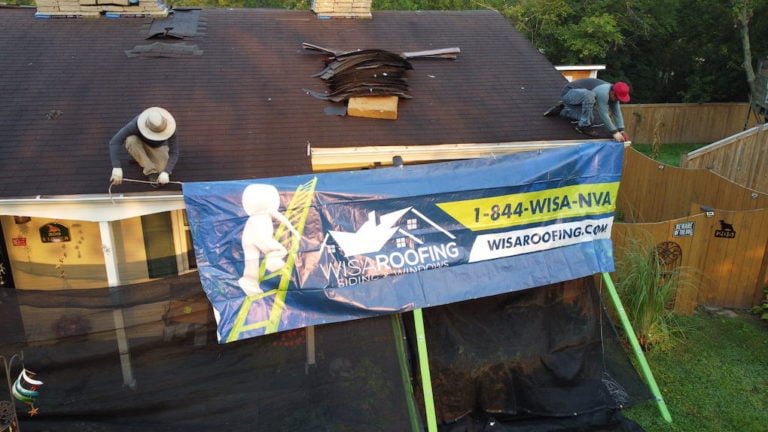 Two workers are installing a large banner on the roof of a house. The banner displays "WISA Roofing Siding Windows" with a phone number and website. The roof appears to be under construction, with tools and materials nearby.