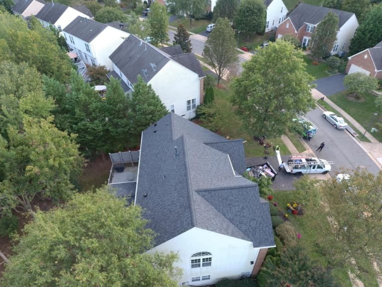 Aerial view of a suburban neighborhood with several detached houses surrounded by trees. A van and a few people are gathered on a driveway beside one of the houses. The scene captures a peaceful residential area.