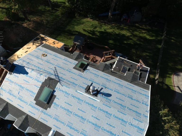 Aerial view of a house roof under renovation, covered with blue CertainTeed underlayment. Two skylights are visible, and materials are scattered around. Nearby, a wooden deck and lush green trees can be seen.