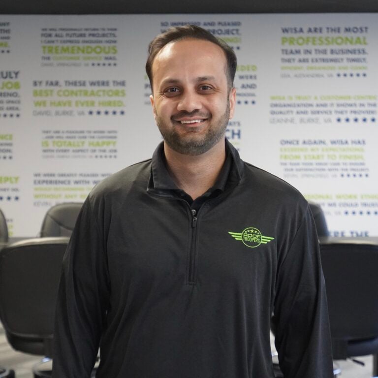 A smiling man with short hair and a beard stands in a conference room, wearing a black zip-up shirt with a green logo. Behind him, a wall is covered with large positive review quotes in blue and green text.