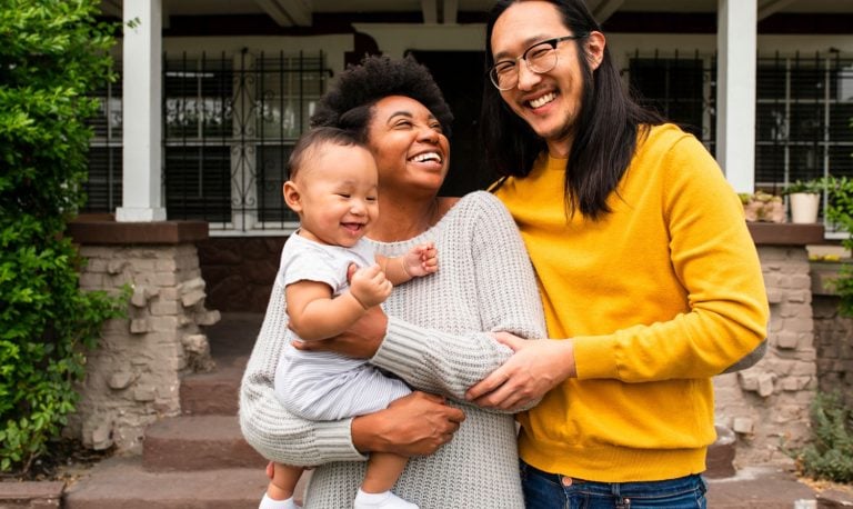 A joyful couple stands on a porch with their baby. The woman holds the baby, smiling widely, while the man beside her beams happily, placing a hand on her shoulder. The background shows a house with windows and a partial view of greenery.