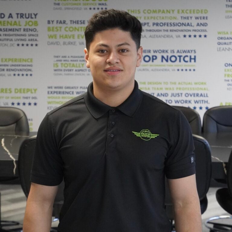 A young man in a black collared shirt stands and smiles in a conference room with motivational quotes covering the wall behind him.
