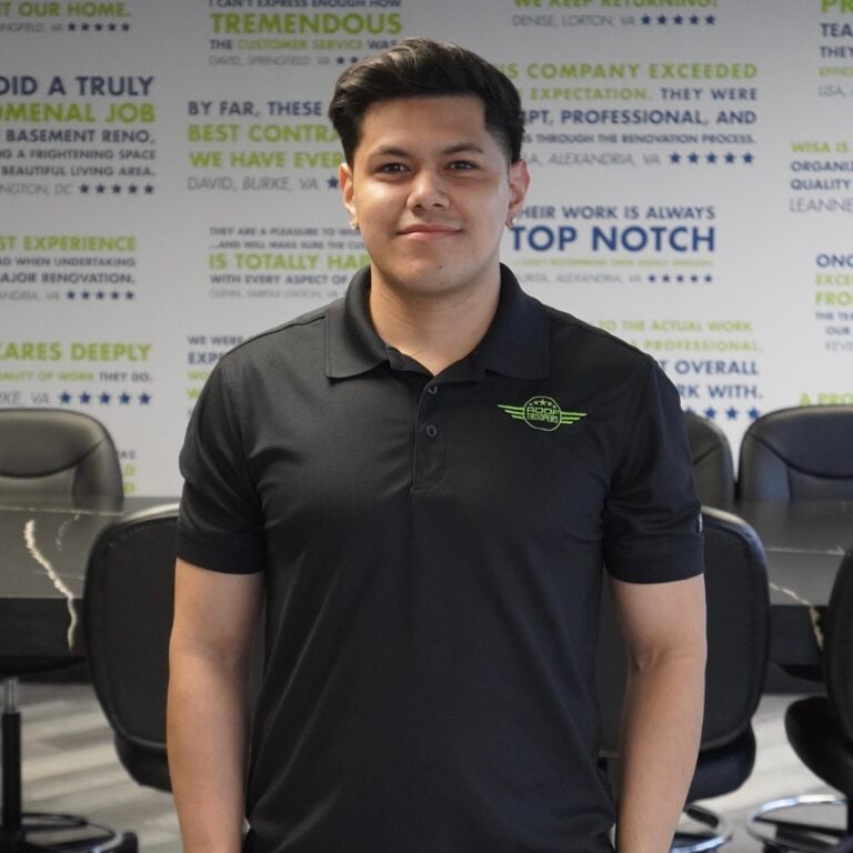 A young man with short dark hair wearing a black polo shirt stands smiling in a modern office with motivational quotes on the wall behind him and conference chairs in the background.