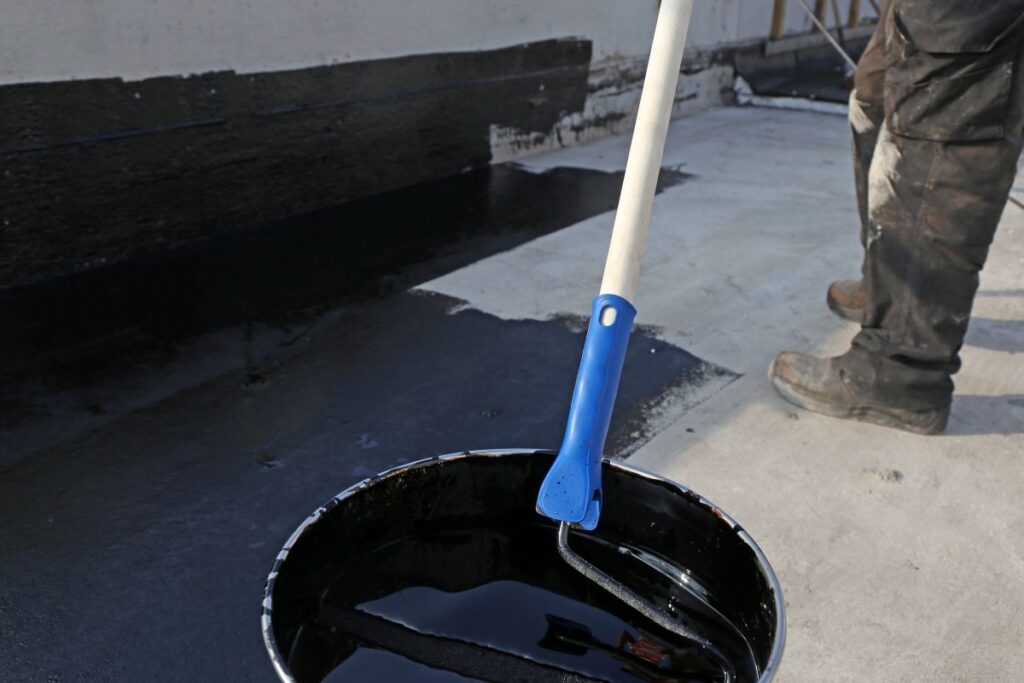 A paint roller with a blue handle is dipped in black waterproofing liquid, as a person applies the best roof sealant for leaks to a concrete surface nearby.