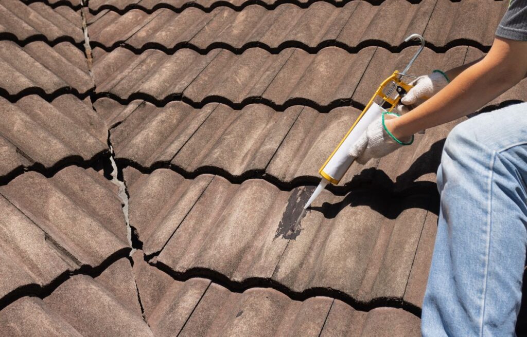A person wearing gloves uses a caulking gun and one of the best roof sealants for leaks to fill a crack in brown roof tiles while kneeling on the wavy-patterned rooftop. The large crack is being carefully repaired.