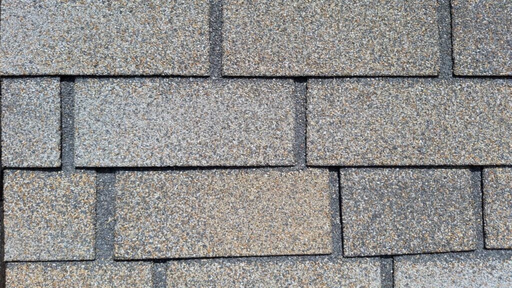 Close-up view of gray asphalt shingles on a flat roof, arranged in an overlapping pattern and showing the texture and detail of the roofing material.