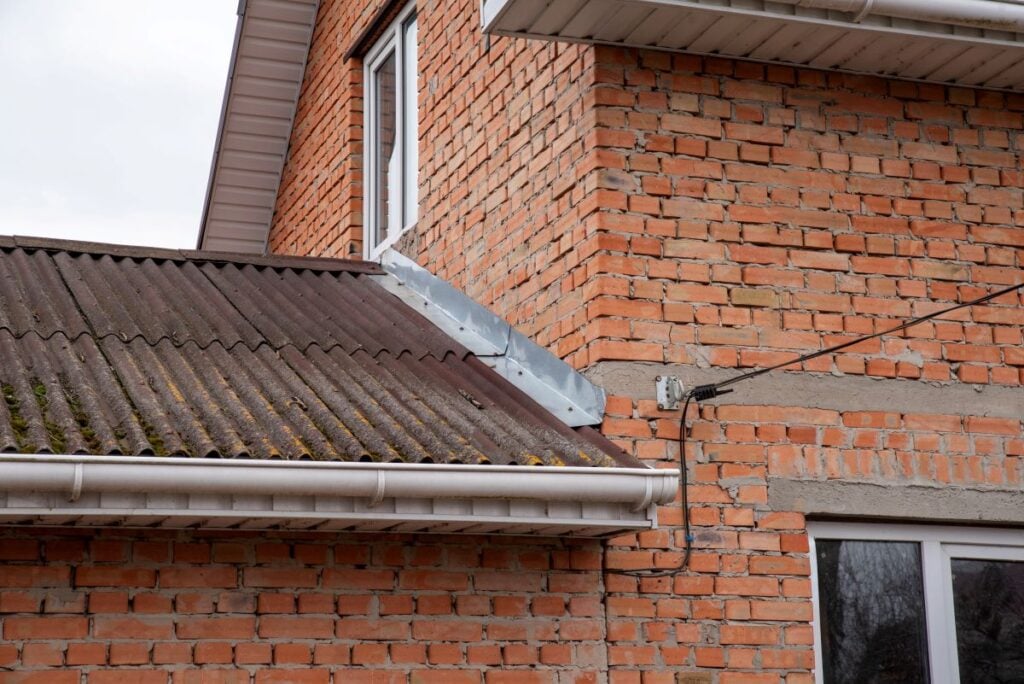 Brick house with upper and lower windows, a corrugated metal roof featuring visible roof flashing repair, leaf debris on shingles, a white rain gutter, and exposed electrical wires attached to the exterior wall.