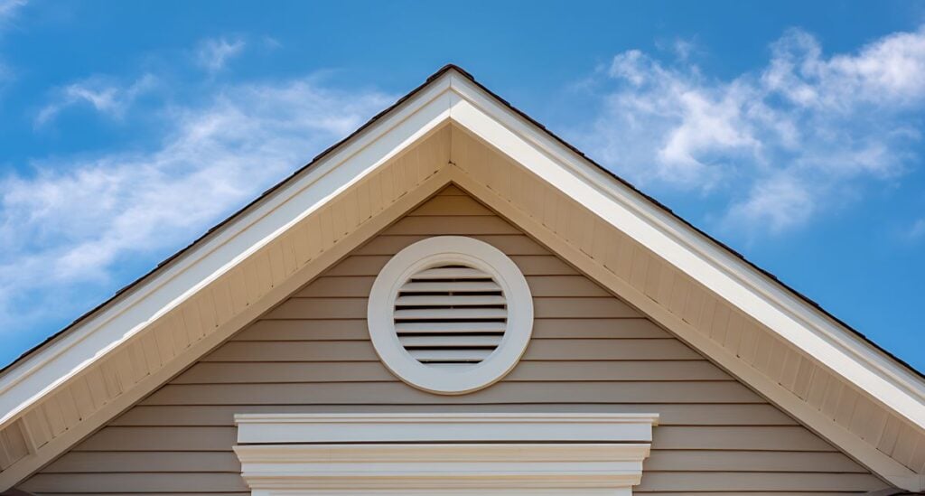 The image shows the upper section of a house with beige siding, a white circular attic vent, decorative trim, and a prominent rake roof overhang under a gabled roof, set against a blue sky with scattered clouds.