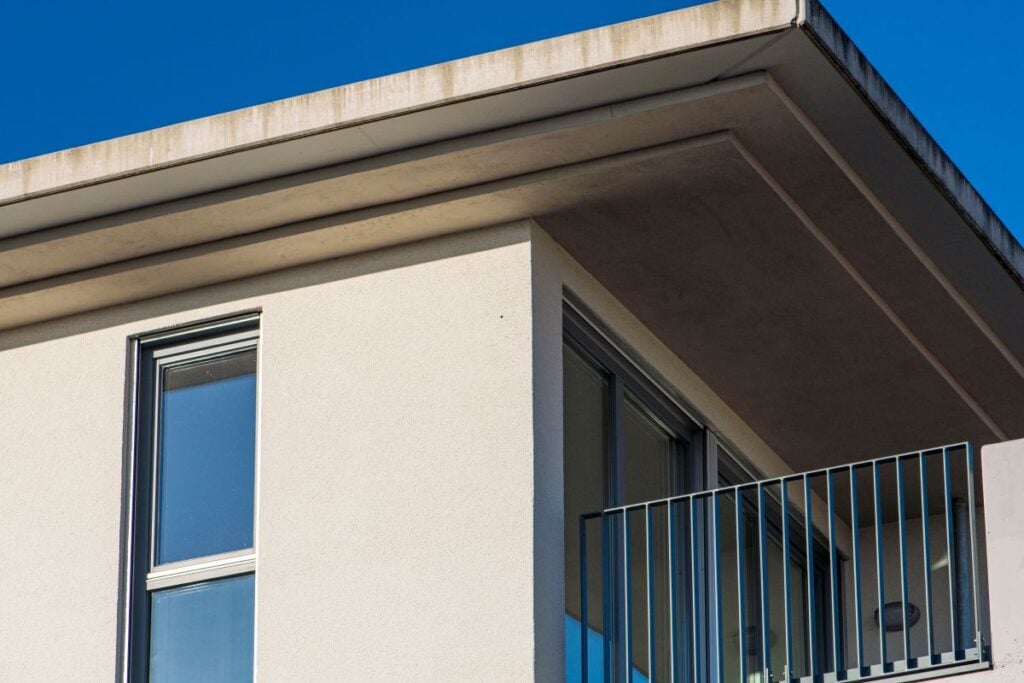 A close-up view of a modern building corner with large windows, a small balcony with metal railings, and a flat roof featuring a sleek roof overhang against a clear blue sky.