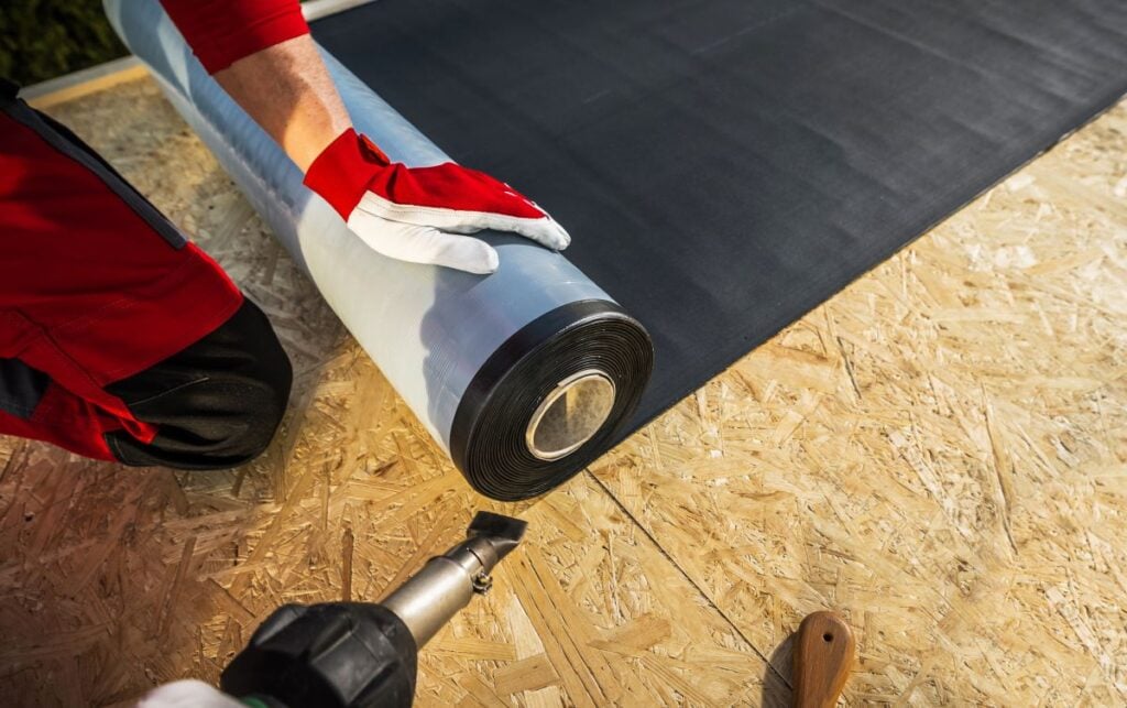 A person in red gloves unrolls roofing material onto a wooden surface, preparing to apply it. A heat gun and a wooden tool are placed nearby on the work surface, suggesting attention to detail and quality—important factors when considering rubber roof cost.
