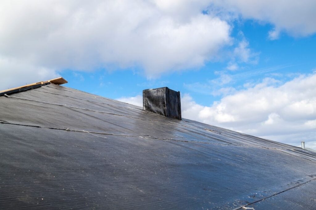A close-up view of a rooftop covered in black waterproof material with a small black chimney, under a partly cloudy blue sky, highlights the sleek finish often associated with modern installations and hints at the affordability of rubber roof cost.