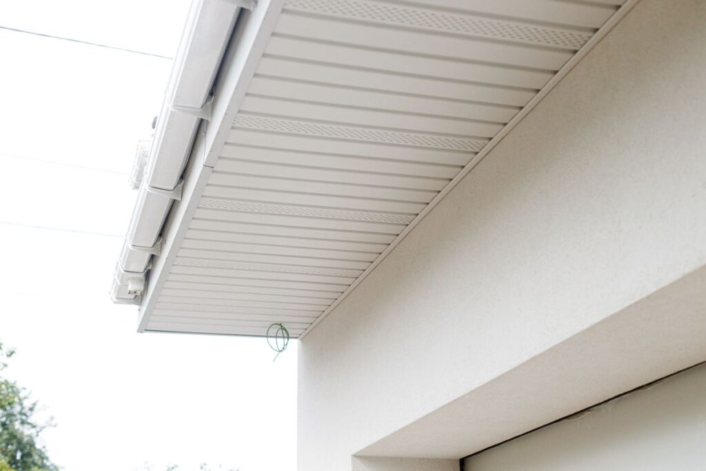 Close-up view of a house’s roof overhang, showing white ventilated soffit and fascia panels under the roofline, part of the gutter, and a small green hook attached near the edge.