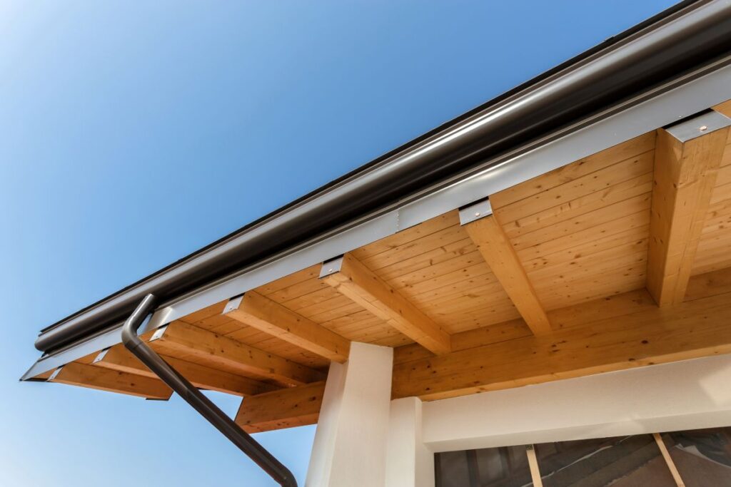 Close-up view of a house roof with exposed wooden beams, a metal gutter, and a downspout beneath the roof overhang against a clear blue sky.