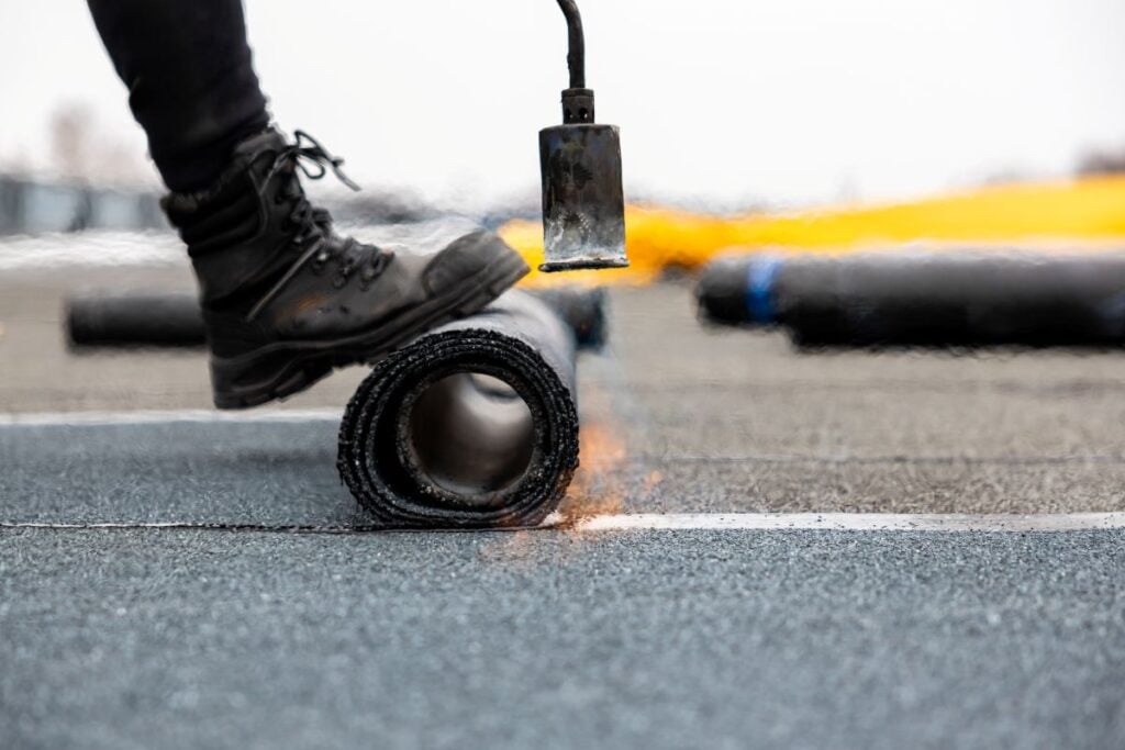 A worker uses a torch to heat and install a rolled roofing material on a flat rooftop, demonstrating a common method for keeping rubber roof cost affordable. The worker wears black boots, and the surface is dark and textured.