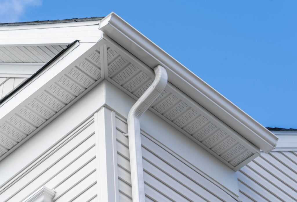 Close-up view of a house's white vinyl siding, roof overhang, soffit, and gutter system with a blue sky in the background. The gutter runs along the roof edge and downspout extends vertically down the side.