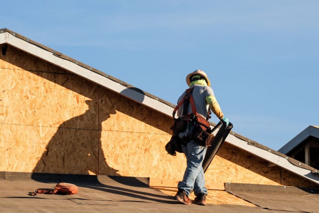 roof financing worker wearing safety gear and a helmet stands on a roof, holding building materials. Plywood sheathing is visible, and the worker’s shadow is cast on the unfinished house under a clear blue sky—perfect for discussing roof financing options.