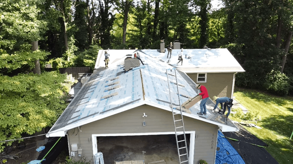 Several workers are installing roofing materials on a house surrounded by trees, demonstrating how to replace missing shingles. The roof is partially covered with underlayment, and ladders lean against the house as the team works on different sections.