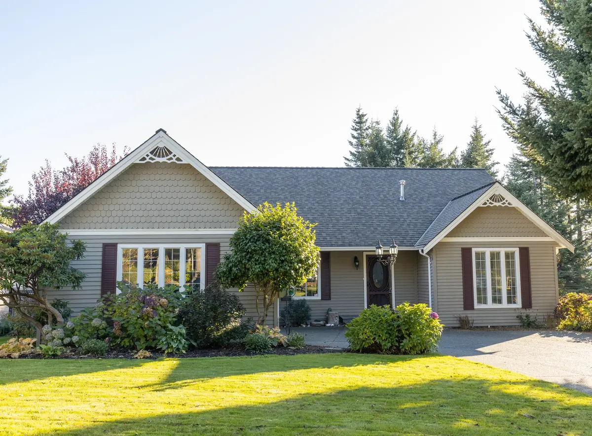 A single-story house with gray siding, white trim, and brown shutters is surrounded by trees and shrubs, with a well-maintained green lawn in front. Under the clear, sunny sky, it's an ideal spot to learn how to replace missing shingles.