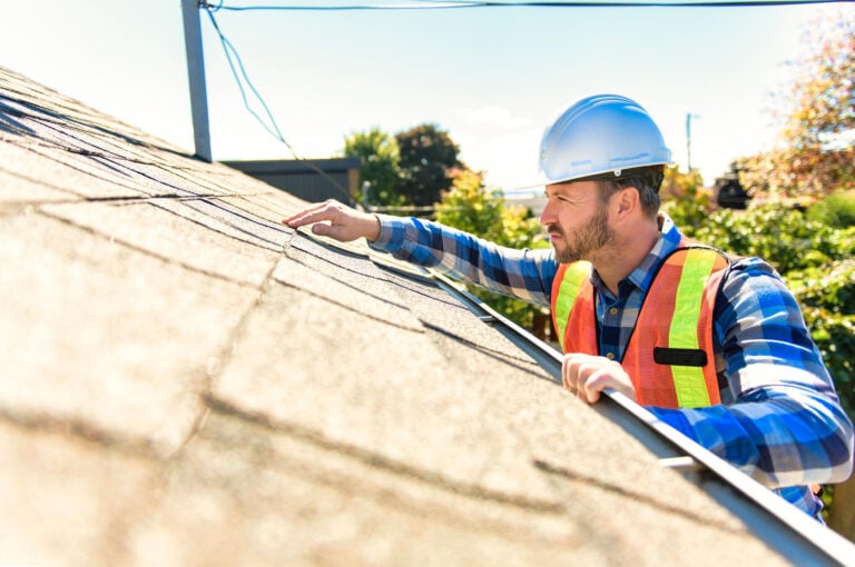 how long does a roof inspection take-man in safety gear stands on a ladder inspecting edge of roof and shingles
