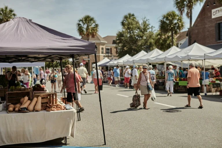 An outdoor farmer's market in Springfield, VA offers a delightful escape, with people browsing stalls under white tents. Vegetables like squash and potatoes are displayed on tables, while palm trees and buildings grace the backdrop under a clear sky—a perfect thing to do in Springfield.