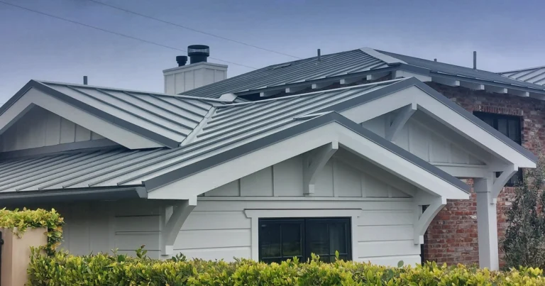 House with a modern gray metal roof, white siding, and brick chimney. The structure features multiple gables and black-framed windows, surrounded by lush greenery.