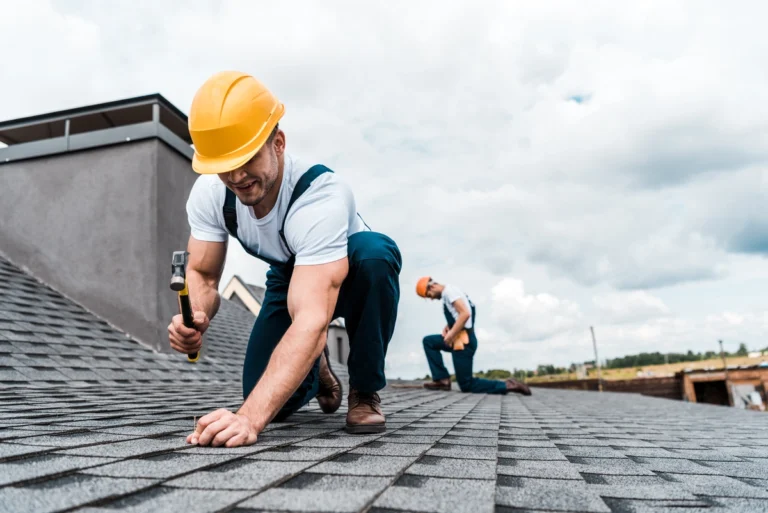 Two construction workers in yellow hard hats and overalls are working on a shingled roof. One is using a hammer, while the other checks shingles. The sky is cloudy in the background.