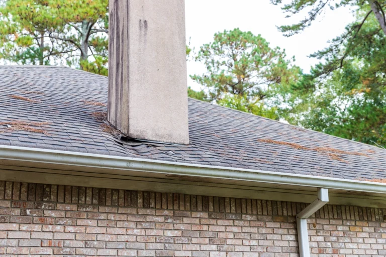 A brick house with a sloped shingle roof and a chimney. The roof is covered with some pine needles, and trees are visible in the background. The image shows a section of white gutter attached to the roof edge.