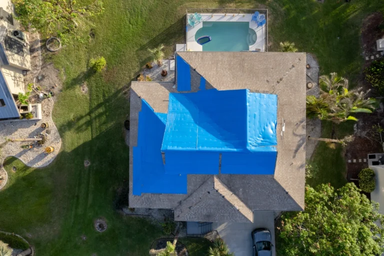 Aerial view of a house with a roof partially covered in blue tarps. The house is surrounded by greenery, and a small rectangular pool is visible in the backyard. A car is parked in the driveway.