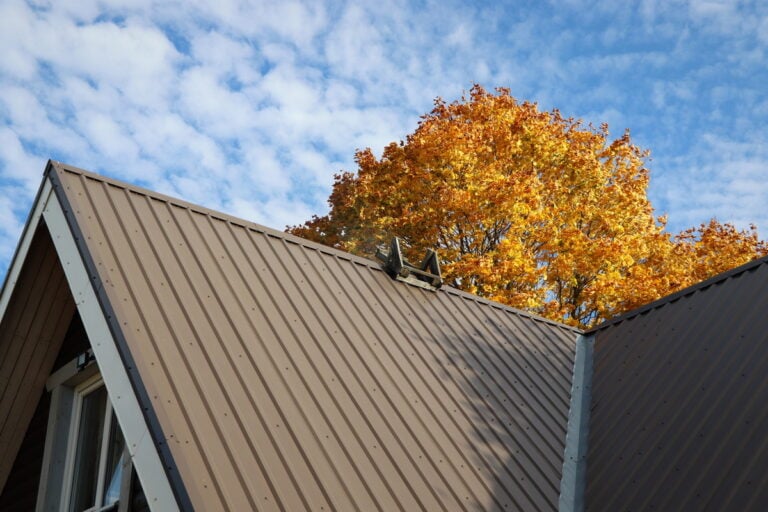 A brown metal roof with a ladder leading to the peak offers both style and durability, lasting decades. In the background, an autumn tree with vibrant orange leaves contrasts against a sky filled with scattered white clouds.