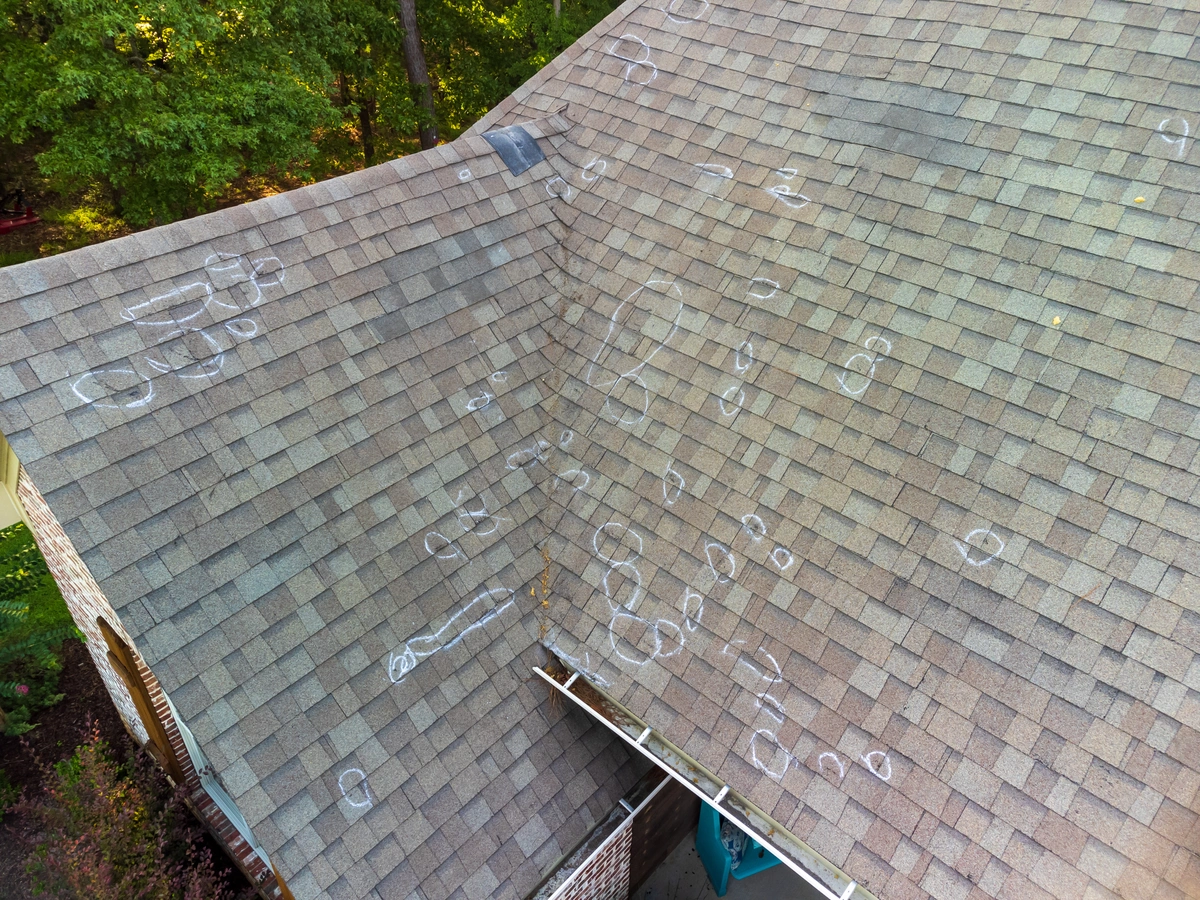 Aerial view of a house roof with numerous chalk circles marking hail damage, hinting at the need for an average insurance payout. The roof shingles are visibly marked, revealing impact points. Surrounding the house are trees and foliage.