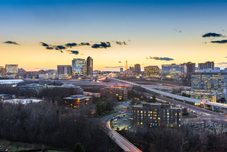 Aerial view of a cityscape at dusk, with illuminated buildings under a gradient sky transitioning from orange to dark blue. Highways curve through the scene, surrounded by leafless trees and headlights from moving vehicles.
