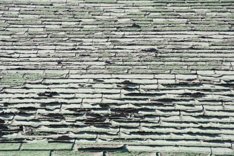 A close-up of an old, weathered shingle roof with peeling surface and visible signs of damage. The shingles appear worn and uneven, with patches of darker material exposed, indicating the need for maintenance or repair.