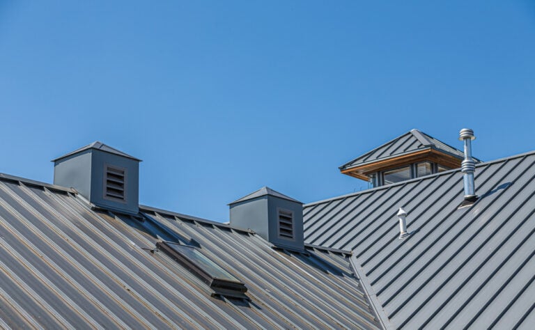 A metal roof with two small vented structures and a chimney against a clear blue sky.