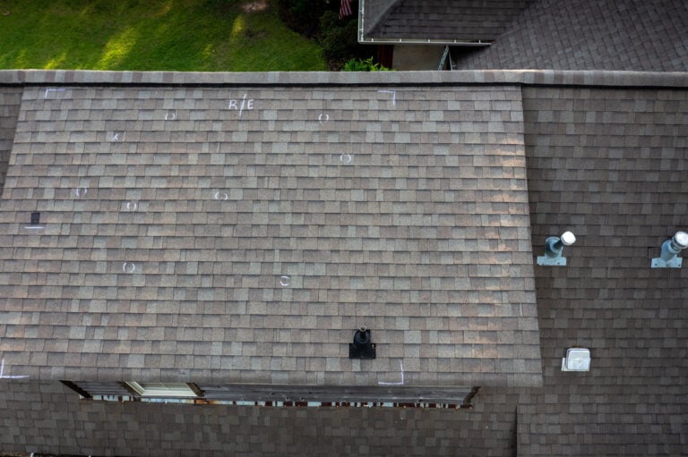 Aerial view of a gray shingle roof with several marked circles indicating areas of concern. Two pipes and a vent are visible on the roof. A grassy yard and part of a neighboring roof are in the background.