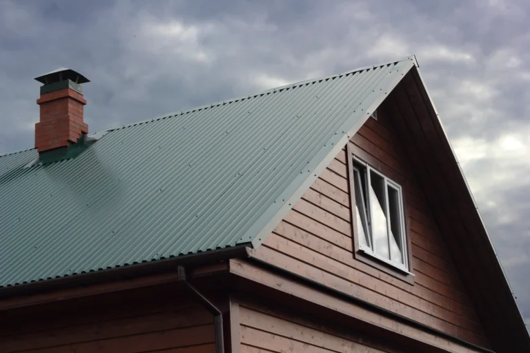 A wooden house with a green metal roof secured by screws on the flat surface and a brick chimney set against a cloudy sky. The roof, with its steep pitch, extends gracefully over a window on the side of the house.