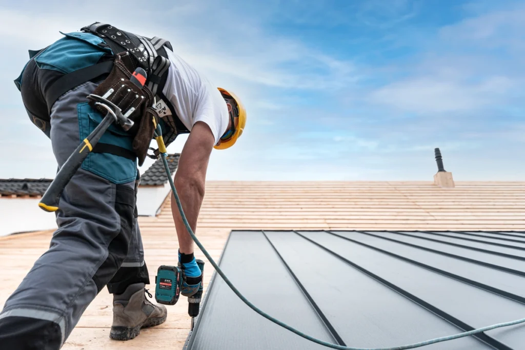Under a clear blue sky, a worker wearing safety gear and a tool belt expertly uses a power drill to install metal roofing panels. Thinking of hiring? Be sure to know the essential questions to ask a roofer before starting your home improvement project.
