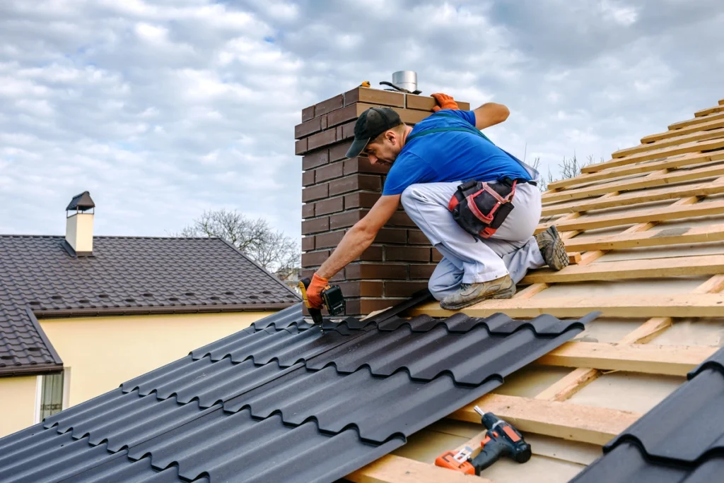 A worker installs black metal roofing sheets on a sloped roof, kneeling by a brick chimney. In his blue shirt, white pants, and orange gloves, he skillfully navigates the task with a nearby toolbox. The scene prompts thoughts of questions to ask a roofer before hiring. A cloudy sky and other rooftops form the backdrop.