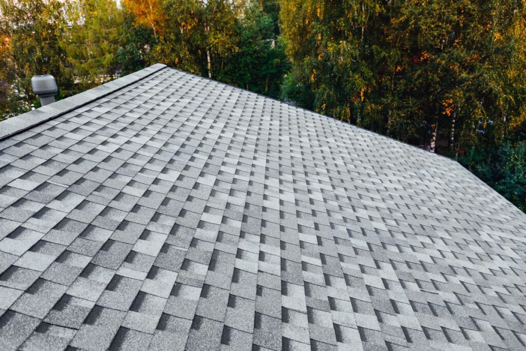 Gray asphalt shingle roof with a geometric pattern, viewed from above, surrounded by green trees with some yellowing leaves; some new roof shingles not laying flat are visible in the arrangement.