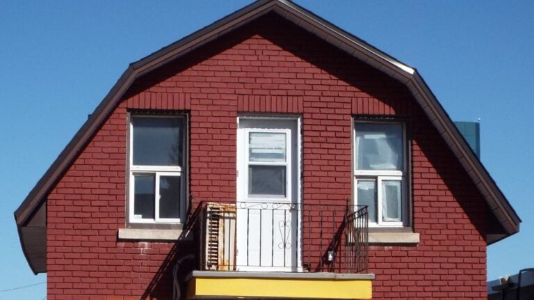 A red brick house with a classic gambrel roof features two white-framed windows and a white door leading to a small balcony with black railings. The sky is clear and blue.