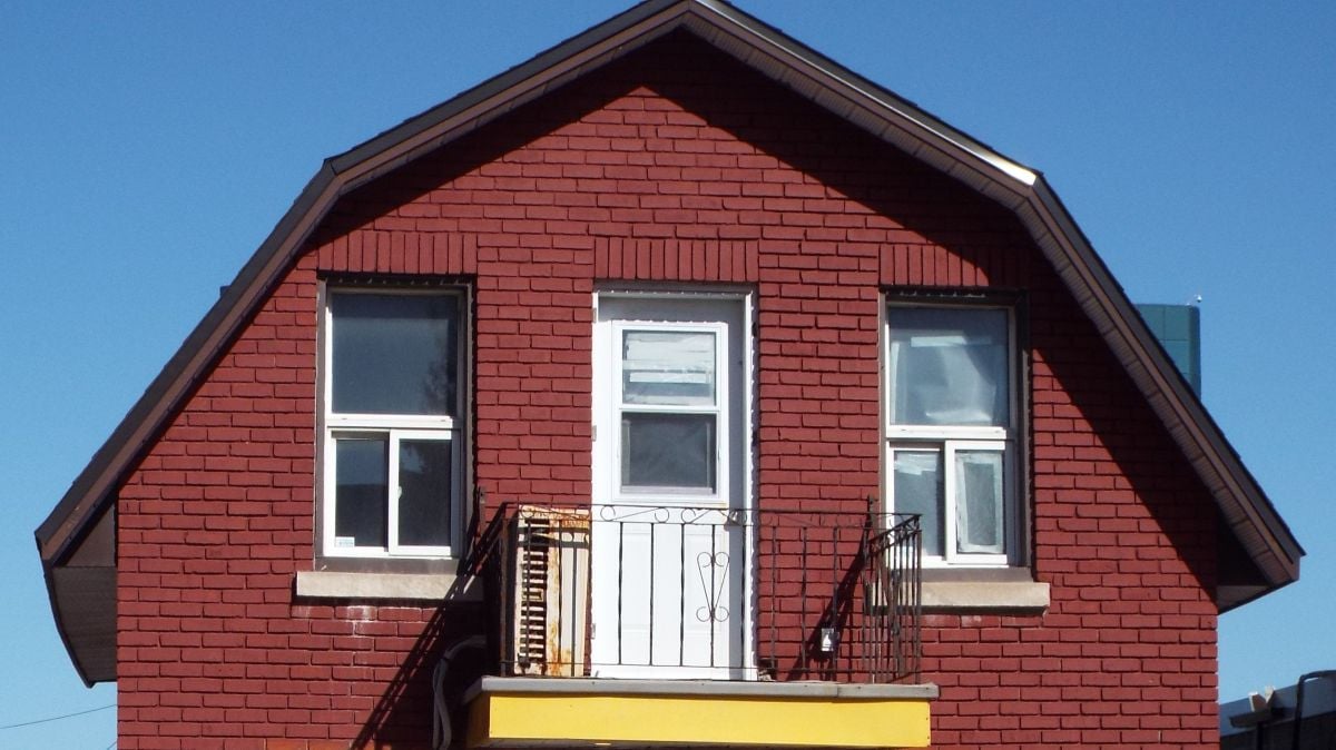 A red brick house with a classic gambrel roof features two white-framed windows and a white door leading to a small balcony with black railings. The sky is clear and blue.